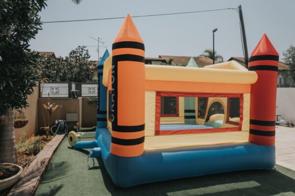 א מתקן קפיצה מתנפח גדול | D4234 | צבעי פנדה של Jumpy Jump עם משטח החלקה וקפיצה, המעוצבת כמו טירה צבעונית עם שני מגדלים מחודדים, מוקמה בחצר דשא. זה בעיקר צהוב עם מבטאים אדומים וכחולים, מוקף בעצים, אזורי בטון ומבנים ברקע.