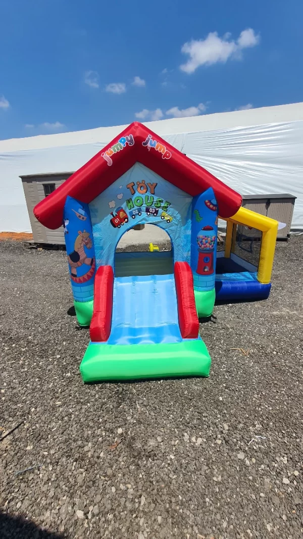 טירה מתנפחת מתנפחת Jumpy Jump הכוללת דמויות מצוירות ו"בית הצעצוע" למעלה, עם מגלשה קדמית ובור כדורים, על רקע שמיים כחולים צלולים על חצץ.