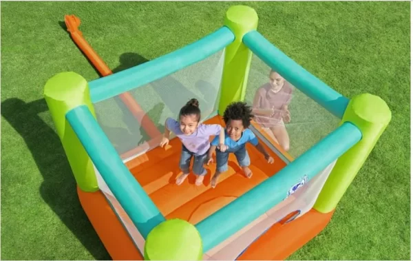 שני ילדים קופצים בשמחה ב-53394bestway-soar מתקן קפיצה הצבעוני על מדשאה עשבונית. מבוגר צופה בהם בפנים. הבית המקפיץ מתהדר בצבעי ירוק וכתום עם ציפוי רשת.